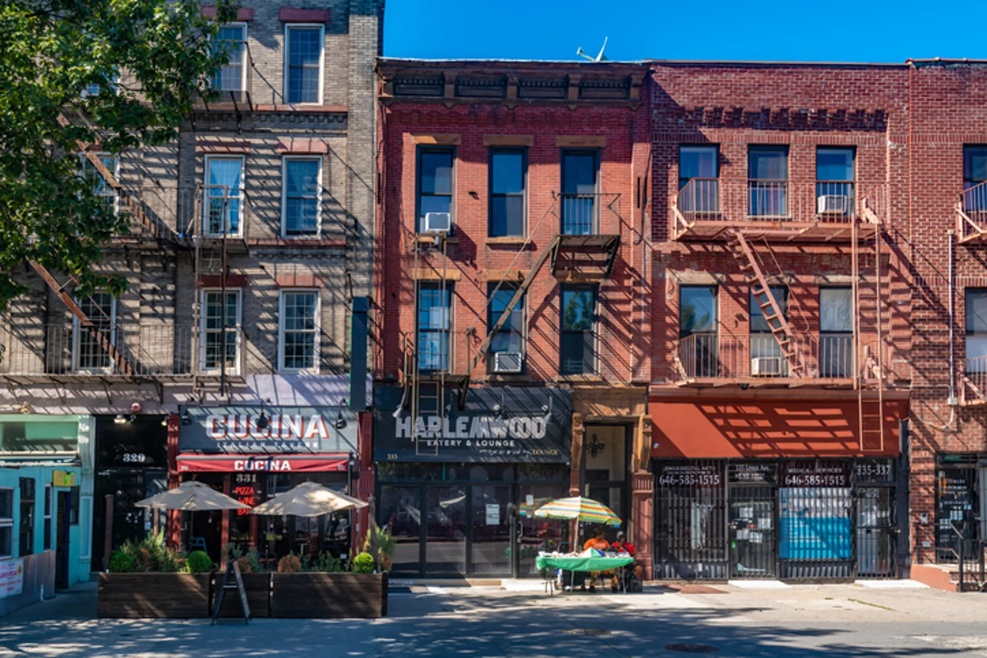 A row of historic brick buildings with ground-floor commercial spaces is seen along a street in Harlem neighborhood