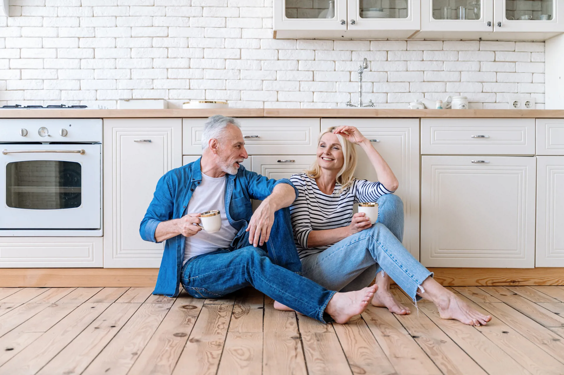 Smiling mid aged woman and man sit on wooden floor in cozy kitchen
