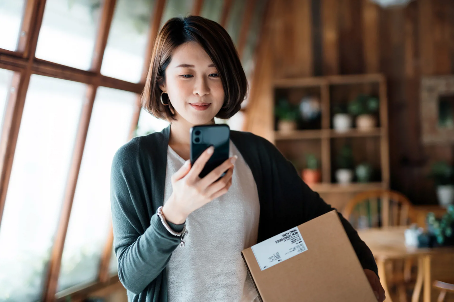 Smiling young Asian woman shopping online with smartphone on hand, receiving a delivered parcel by home delivery service