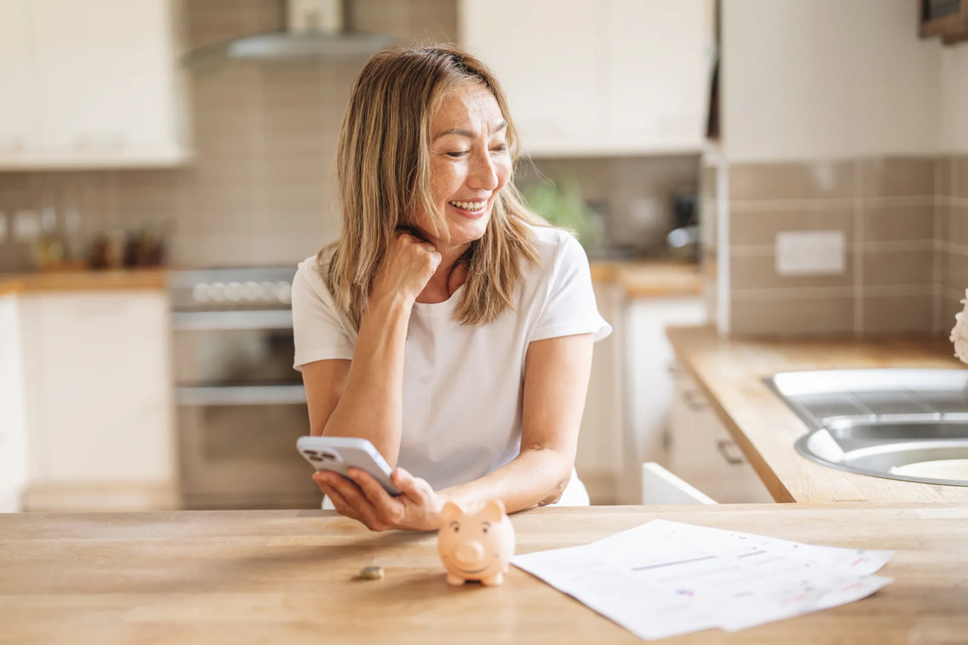 Woman smiling while managing finances at home using smartphone, papers, and piggy bank on kitchen table during daytime